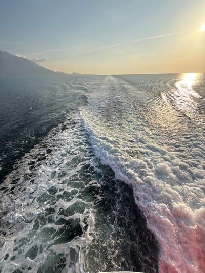 The image depicts a seascape with a vessel's wake trailing behind it in the waters near Palaiopoli, Greece. In the foreground, turbulent white foam and dark blue-green agitated water form the immediate wake, extending diagonally from the lower left towards the upper right. A reddish tint is observed within the foam on the far right of the immediate wake. The wake gradually diminishes into a series of ripples on the water's surface towards the distant horizon. On the far left, a mountainous coastline is visible against a light blue sky, which transitions to a pale yellow and orange hue towards the right, where a brilliant sun is positioned. The sun's reflection creates a bright, shimmering path across the water's surface, intersecting with the distant wake pattern. The sky above is clear, with a faint linear contrail visible in the upper center. No people or specific objects other than the water and distant land are discernible. The scene captures the movement of a boat on open water during what appears to be either sunrise or sunset.