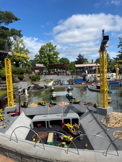 The image depicts a miniature construction site scene in Billund, Denmark, likely at LEGOLAND Billund. Towering yellow construction cranes are prominent features, one on the left and another on the right. The scene includes a body of water with several tugboats and construction vehicles like excavators and dump trucks, all rendered in LEGO bricks. A small bridge arches over a section of the water.

The environment features lush green trees and a partly cloudy blue sky. In the background, there are shaded seating areas where numerous LEGO minifigures are gathered, suggesting a public attraction. The ground is a mix of gray gravel, dark gray surfaces, and piles of brown and tan LEGO bricks representing construction materials. Several LEGO minifigures are actively engaged in construction activities around the site, operating machinery and moving materials. The foreground shows a concrete barrier with curved metal railings. The overall impression is a detailed and bustling miniature diorama of a construction operation.