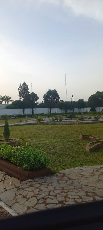 The image depicts an outdoor scene in Barikin Jirga Summa, Nigeria. In the foreground, a pathway paved with irregular flagstones leads into a well-maintained lawn area. To the left, a raised planter bed contains lush green shrubs. Beyond the lawn, a paved walkway winds through the grassy area, bordered by low hedges and ornamental plants. Several streetlights are visible along this walkway. In the background, a white wall with a fence runs horizontally across the image, with various trees and palm trees planted behind it. Two tall communication towers stand prominently against the pale sky, one taller than the other. The overall scene suggests a public park or a well-landscaped area within a city. There are no people visible in the image.