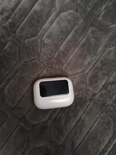 A white charging case for wireless earbuds rests on a dark gray quilted blanket. The case is oriented horizontally, with its screen facing upwards and slightly to the left. The blanket exhibits a pattern of diagonal ridges. The scene is captured from a top-down perspective. There is no indication of the location being Polovinnoye, Russia.