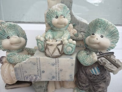Three ceramic hedgehog figurines are positioned on a white surface, likely a windowsill or ledge, in front of a slightly out-of-focus background that appears to be a window with a cloudy sky reflected. The figurines are arranged in a semi-circle.
The leftmost figurine is facing slightly to the right, wearing a cream-colored dress with yellow dots. Its hands are clasped in front of its body.
The central figurine is seated and facing forward, appearing to play a small drum. It has a light blue-green spiky texture on its head and shoulders.
The rightmost figurine is also facing slightly to the right and is holding a violin and bow. It is dressed in dark, baggy clothing with a patched section on its chest featuring blue and white stripes. This figurine also has the characteristic blue-green spiky texture.
The figurines are part of a larger ceramic piece that resembles a piano or keyboard, on which they are seated. This "piano" is decorated with painted lines and circular indentations. The overall color palette of the figurines and the piano is muted, with shades of white, cream, light blue-green, and brown.
The scene is static and appears to be an indoor display. There is no indication of the city of Ceadîr-Lunga, Moldova in the image itself; this contextual information is provided externally.