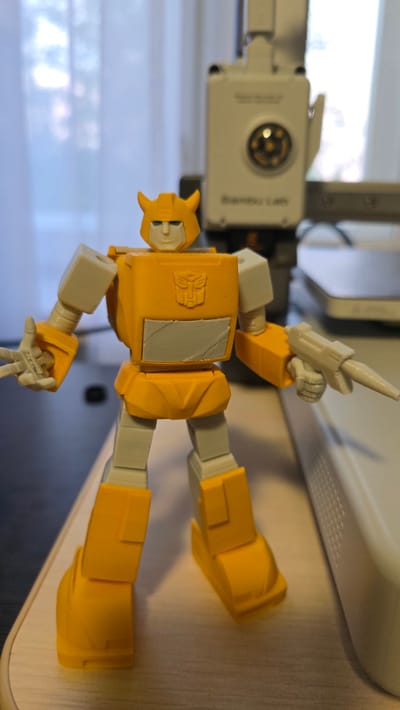 A yellow and white Transformer action figure stands on a wooden surface. The figure is in a humanoid pose with its arms slightly out. Its left hand is held up, with the index and middle fingers extended in a peace sign. Its right hand holds a white blaster accessory. The figure has a yellow helmet with horns, a white face with blue eyes, a yellow chest with a gray Autobot insignia, and yellow legs. In the background, a white and gray machine with a circular lens is visible, as is a blurred image of what appears to be a window with curtains. The scene is lit with soft, artificial light. The image is taken from a slightly low angle, looking up at the action figure.