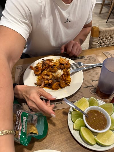 A table setting features a plate of grilled shrimp with rice, accompanied by a plate of lime wedges and a small dish of salsa. A blue-tinted drinking glass and a bag of toasted corn tortillas are also present. A diner wearing a white t-shirt with a logo sits at the table, holding a fork and knife, ready to eat. A menu with Spanish text listing various shrimp dishes is visible in the background.