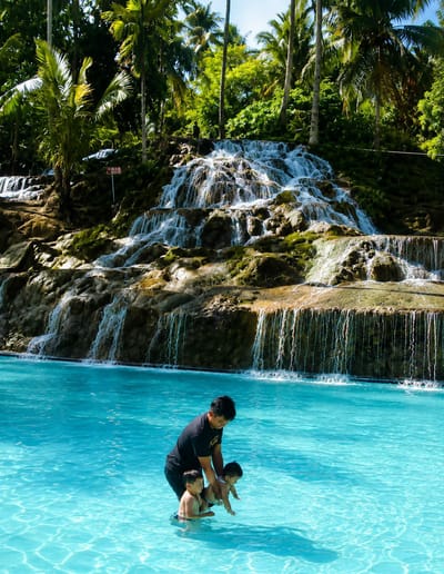 An adult male wearing a black t-shirt and dark shorts is in a bright turquoise swimming pool, holding two young children. The man is positioned to the left of the frame, facing towards the right. He is holding one child, a baby, with both hands, seemingly lowering it into the water. Another child, a boy wearing only swim trunks, is standing next to the man, to his left. The boy appears to be reaching towards the baby. The pool has clear, rippling water, reflecting the sunlight.
Behind the pool, a multi-tiered waterfall cascades down a rocky cliff face covered in moss and greenery. Palm trees and lush vegetation form a dense backdrop, with some trees reaching tall into the sky. A sign with red lettering is visible on the left side of the waterfall, partially obscured by foliage. The scene appears to be a natural attraction, possibly a resort or a park, featuring the waterfall and the swimming pool. The location is identified as Silaiyajae, Indonesia.