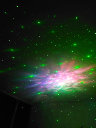 The image depicts a ceiling projected with green laser dots resembling stars, interspersed with a colorful nebula-like cloud of pink and purple light. A dark, rectangular area, possibly a doorway or window, is visible in the lower left portion of the frame. The scene is devoid of people or any discernible activity. The location in Areni, Armenia, is not ascertainable from the visual information provided.