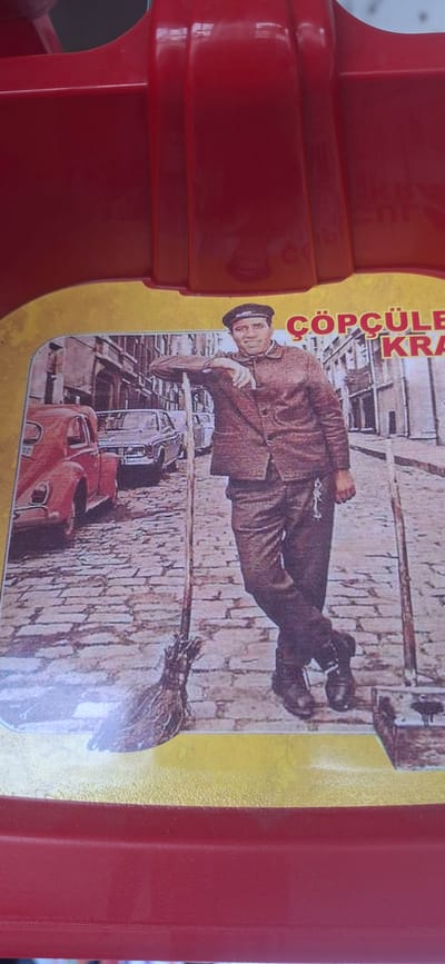 The image shows a framed picture on a red surface. The picture depicts a man in a brown suit and cap leaning on a broomstick, with his legs crossed. He is standing on a cobblestone street in a city, with buildings in the background and cars on the left. The man is smiling and looking towards the viewer. The text "ÇÖPCÜLE" and "KRA" is visible above the man's head. The red surface appears to be part of a toy or a container. There is no indication of the location being Zugdidi, Georgia.