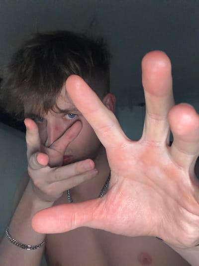 The image shows a close-up, frontal view of a young person with brown hair and blue eyes. The subject's face is partially obscured by their hands, which are positioned in front of their face. The person is wearing a silver chain necklace and a silver bracelet on their left wrist. The background is dark and out of focus, suggesting an indoor setting with low lighting. The image does not contain any visual information related to the city of Vanino, Russia.