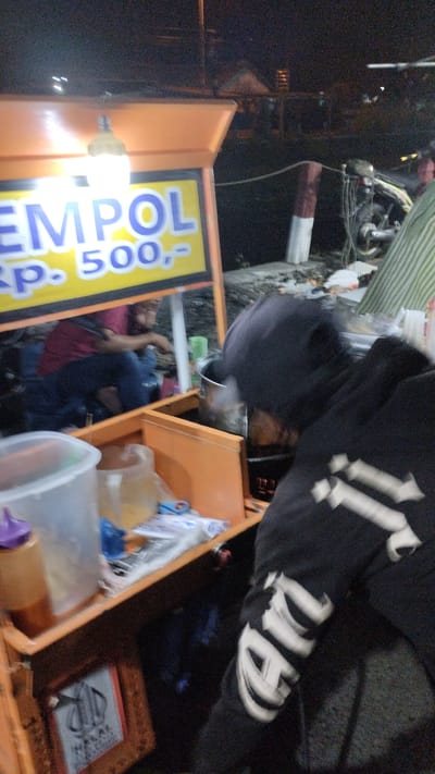 The image depicts a nighttime scene of a street food cart in Sukodono, Indonesia. The cart is orange and yellow, with a sign displaying "EMPUL RP. 500,-". A person wearing a dark hoodie with white lettering on the sleeve is partially visible in the foreground, facing away from the camera. Inside the cart, a person in a maroon shirt is seated. Various containers, including a plastic one with a purple lid, are visible on the cart. In the background, a motorcycle is parked, and a striped awning is visible. The environment appears to be an outdoor, possibly urban, setting.