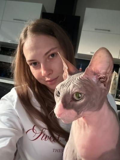 The image captures a close-up, selfie-style shot of a young woman and a Sphynx cat. The woman, with long, light brown hair and light eyes, is wearing a white sweatshirt with dark red lettering that partially reads "Sweet" and "THES". She is looking towards the camera with a soft smile. The Sphynx cat, with its characteristic hairless, wrinkled skin in shades of gray and pink, is positioned to the right of the woman, looking slightly off-camera. Its large ears are prominent. The background appears to be an indoor domestic setting, possibly a kitchen, with white cabinets visible. The lighting suggests an indoor environment, perhaps during the evening or with artificial lighting. No specific details definitively place the image in Novaya Ivanovka, Russia, but it depicts a common domestic scene.