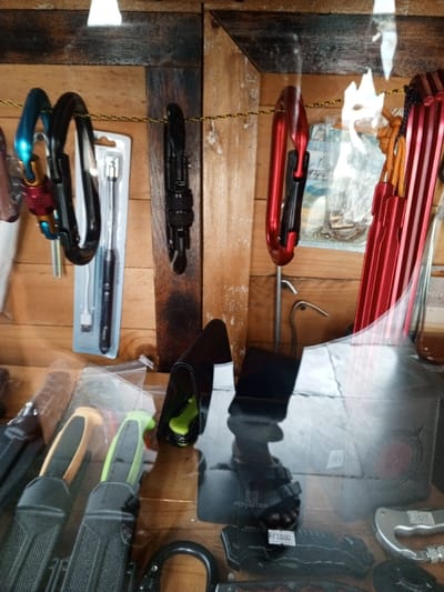 The scene depicts a close-up view of a retail display for outdoor and utility equipment, located within a store or stall in Baza, Myanmar. The background is a wall constructed from varied wooden planks, arranged both vertically and horizontally. A yellow and black braided cord is strung across the upper mid-section of this wooden backdrop.
Hanging from the braided cord on the left are a blue and red carabiner and a black carabiner. Behind them, a packaged item, possibly a tool, is visible in a clear plastic blister pack. A black multi-tool or tactical pen-like object hangs from the cord in the center, while multiple red carabiners, some stacked, are suspended on the right.
The lower half of the image shows items behind a reflective glass surface. Visible objects include several knives or tools in black sheaths, with yellow and green handles partially exposed. A black holster containing a bright green item is present, along with other dark-colored items. A prominent reflection of a human foot wearing a dark sandal is visible on the glass in the lower middle to right section. A white price tag displaying "Rp 70000" is attached to a black item in the lower right, next to a partially visible silver carabiner. No direct human interaction or explicit actions are observed beyond the static display of merchandise and the reflection.