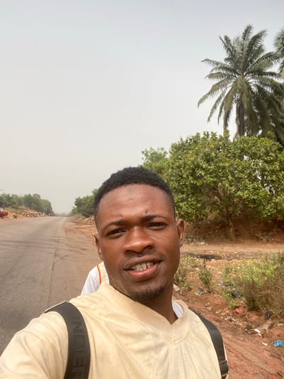 The image captures a male individual in the foreground, facing the camera with a slight smile, positioned in Okura, Nigeria. He has short dark hair and a light beard, wearing a light-colored long-sleeved top with a black backpack strap visible over his right shoulder. A second individual is partially obscured behind him to the left, visible wearing a white shirt with a colorful pattern near the collar. The background features a two-lane asphalt road extending into the distance on the left, with a red vehicle partially visible further down the road. To the right of the road, the terrain consists of unpaved, reddish-brown earth with scattered debris and low-lying vegetation. The right side of the frame and the mid-ground are dominated by dense green foliage, including several tall palm trees and various bushes. The sky overhead is uniformly overcast and hazy.