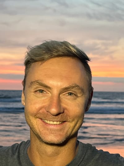 The scene captures an adult male in a close-up, selfie-style portrait, positioned against a coastal backdrop in Kerobokan Kelod, Indonesia. The subject, with fair skin, light-colored eyes, and short, light brown-to-grey hair, is seen from the chest up, wearing a dark grey t-shirt. He displays a smiling expression, revealing upper teeth, with light stubble on his lower face. The background consists of an ocean with visible waves, meeting a horizon line under a sky characterized by sunset or sunrise colors including pale orange, pink, and light blue, interspersed with clouds. No other individuals or significant objects are present within the frame. The action is a self-portrait; no interaction with others is depicted.