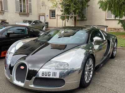 A chrome and black Bugatti Veyron supercar is prominently parked on a paved surface in Molsheim, France. The vehicle features a reflective chrome lower body and fenders, contrasting with a glossy black hood and roof. Its front displays the distinctive Bugatti horseshoe grille with a red oval badge and a front license plate reading "BUG III". Polished chrome multi-spoke wheels are visible. To the immediate left and slightly behind, a second black Bugatti, likely another Veyron, is partially visible, showing an orange accent on its lower side. Further in the background, a third silver-colored classic-style coupe, possibly a Bugatti Type 57, is parked. The backdrop consists of an aged, light-colored building with stucco or stone walls, featuring multiple windows with closed white louvered shutters. A small black letter 'a' is stenciled on the building wall near an entrance. Green foliage from trees is visible on the right side and partially behind the vehicles. The scene is illuminated by natural daylight, casting reflections on the cars' surfaces.
