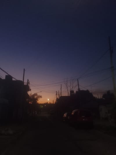 A street scene at dusk, the sky transitioning from deep blue to orange near the horizon. Silhouetted buildings and power lines frame the view down the street, where a faint street light glows. The foreground is dark, with a few parked vehicles visible on the right side. A bare tree and other obscured foliage add to the silhouette effect. The overall mood is peaceful and quiet.