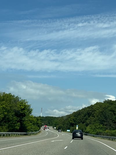 The media file shows a scenic highway curving through a landscape of dense green trees. The road is paved and marked with white lines, with a metal guardrail on the right side. Several vehicles are visible on the highway, including a black car in the foreground and a red and white truck further down the road. In the distance, a tall, white tower stands against a backdrop of clouds and a blue sky. The sky is partly cloudy, with wispy cirrus clouds stretching across the frame. The overall impression is one of a bright, clear day, possibly in the late morning or early afternoon. The image appears to be taken from a moving vehicle, suggesting a journey or travel. The lush greenery and the road winding through the trees evoke a sense of nature and open space. The presence of the tower could indicate a rural or semi-rural setting, perhaps near a town or a communications hub. No people are clearly visible, but the presence of vehicles implies human activity. There are some blue signs with white symbols visible on the side of the road, but the text is not legible. The location is Lermontovo, Russia.