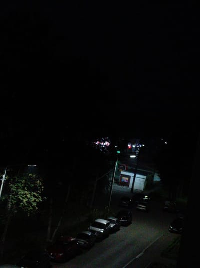 The image depicts a street scene at night, likely in Alatyr, Russia, given the context. A residential building or apartment block is visible in the foreground, with a dark, possibly paved, road stretching out below. Several cars are parked along the road. The street is illuminated by artificial lights, including a streetlight to the left and lights from buildings or traffic in the distance. The overall atmosphere is dark and somewhat desolate, characteristic of a nighttime urban environment. There are no visible people in the image, and the focus is on the infrastructure and vehicles. The vegetation, including trees and bushes, suggests an urban or suburban setting with some green spaces. The quality of the image is grainy, likely due to low light conditions.
