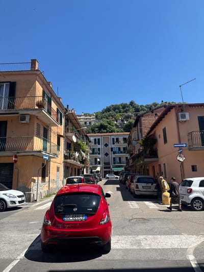 The image shows a narrow street in Trevignano Romano, Italy, lined with old buildings and parked cars. A red Lancia Ypsilon is prominently positioned in the foreground, facing away from the camera. Two elderly individuals are crossing the street on a crosswalk near a white car. Buildings on either side of the street are multi-storied with balconies and shutters. A wooded hill is visible in the distance. The sky is clear and blue. A street sign indicating direction and a convex mirror for traffic visibility are affixed to a building on the right.