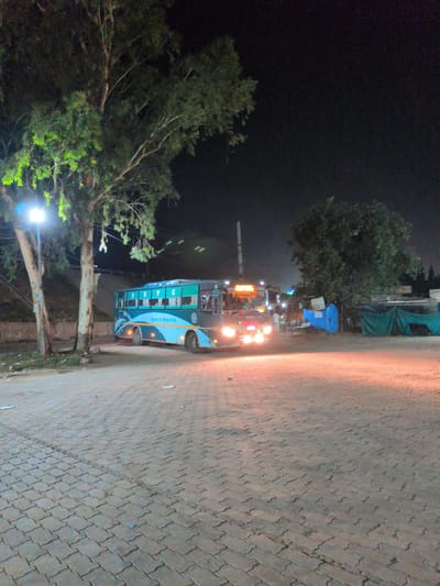 In Samana Bahu, India, at night, a teal and light blue bus with the letters "HRTC" visible on its side is parked on a brick-paved lot. The bus has its headlights on, illuminating the ground in front of it. Two trees stand to the left of the bus, one of which has a bright light fixture attached to its trunk. In the background, there are some structures covered with tarpaulin and more trees. The sky is dark, indicating it is nighttime.