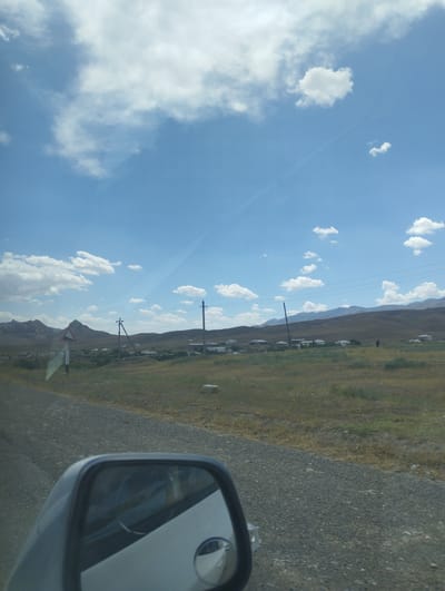 The image depicts a rural landscape with a road in the foreground. A car's side mirror is visible on the left, reflecting a portion of the interior. The road appears to be unpaved, with gravel and some dry vegetation on the sides. In the middle ground, there is a small village with scattered houses and some utility poles with overhead wires. A triangular road sign is visible on the left, indicating a potential hazard or intersection. The background consists of rolling hills and mountains under a bright blue sky with scattered white clouds. The scene suggests a sunny day in a relatively undeveloped area. The location is identified as Asmansay, Uzbekistan.