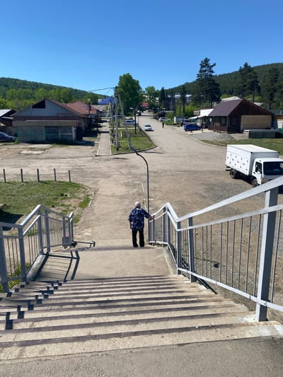 An elderly person wearing a dark blue shirt with a white pattern and black pants walks down a concrete outdoor staircase with metal railings. The staircase leads to a dirt road in the village of Bolshoy Lug, Russia. Along the road are several low-rise buildings with various roof colors, some appearing to be shops or houses. Trees and a forested hillside form the backdrop under a clear blue sky. A white box truck is parked on the right side of the road.