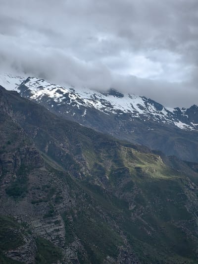The image depicts a mountainous landscape under a cloudy sky, suggestive of a cool, possibly overcast day. The foreground features steep, rocky slopes partially covered in low-lying vegetation. Further into the background, a higher mountain range is visible, its peaks dusted with snow. The clouds are dense and layered, obscuring parts of the sky and casting a diffused light on the scene.
The terrain is rugged and uneven, with visible rock formations and scree slopes. Patches of green vegetation, appearing as grass or low shrubs, cling to the mountainsides, especially in the mid-ground where a patch of sunlight breaks through the clouds, illuminating a specific section of the slope. The snow-capped mountains in the distance suggest a high-altitude location. The overall impression is one of a remote and wild natural environment. The time of day is difficult to ascertain precisely due to the cloud cover, but the light suggests it could be morning or late afternoon. There are no people, objects, or text visible in the image. The scene conveys a sense of dramatic natural beauty, perhaps with a hint of the raw power of the mountains. The location context of Gemur, India, aligns with the visual cues of a high-altitude, potentially rugged mountainous region.
