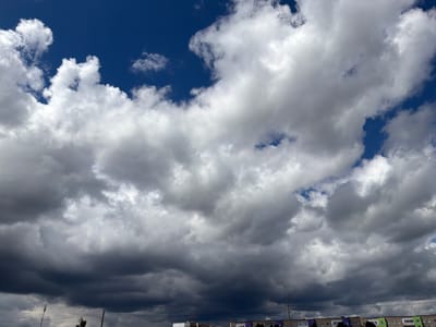 This is a full-frame, eye-level shot of a dramatic sky above the city of Byerazino, Belarus. The sky is predominantly filled with a mix of light and dark cumulus clouds, suggesting an impending storm or a recent one. The clouds are dense and textured, with varying shades of white, gray, and deep charcoal, creating a sense of depth and movement. Patches of deep blue sky are visible through the clouds, particularly in the upper portion of the image. In the lower section of the frame, the tops of multi-story residential buildings are visible, with their rooftops and windows showing. These buildings appear to be of simple, functional design, typical of Soviet-era architecture, and are lined up horizontally across the bottom of the image. The scene is devoid of any people or distinct actions. The overall environment suggests an urban setting on a day with turbulent weather.