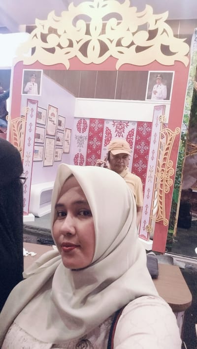 A woman wearing a light beige hijab is taking a selfie in the foreground. She has fair skin and is looking towards the camera with a slight smile. Behind her, a man in a yellow shirt and an orange cap is talking on his phone. He is standing in front of a display with red and white patterned wallpaper. The display also features framed pictures and ornate wooden cutouts. In the background, two framed portraits of men in uniform are visible on the upper part of the display. The overall setting appears to be an indoor exhibition or event in Medan, Indonesia. The lighting suggests it is daytime. There is no visible text in the image.