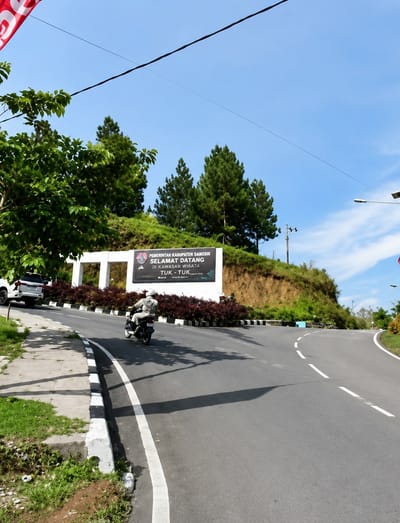 The image captures a view of a road in Turpuk Sihotang, Indonesia, with a white SUV parked on the left side. A person rides a motorcycle on the road, heading away from the camera. To the right of the road, a sign displays text in Indonesian, and beyond that, a hillside covered in vegetation. The sky is clear and blue, with a few scattered clouds. The road curves to the right, leading into the distance. The scene appears to be set in a natural environment, with trees and greenery surrounding the road. The overall atmosphere is peaceful and serene.