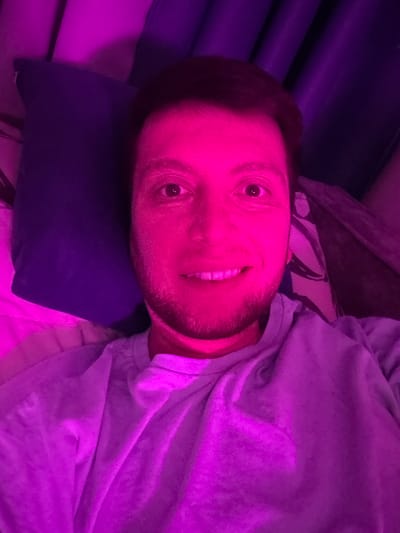 A man is taking a selfie with a purple light. He is lying down on a pillow with a blue pillow behind it. He is wearing a light purple t-shirt. The background is dark and appears to be indoors.