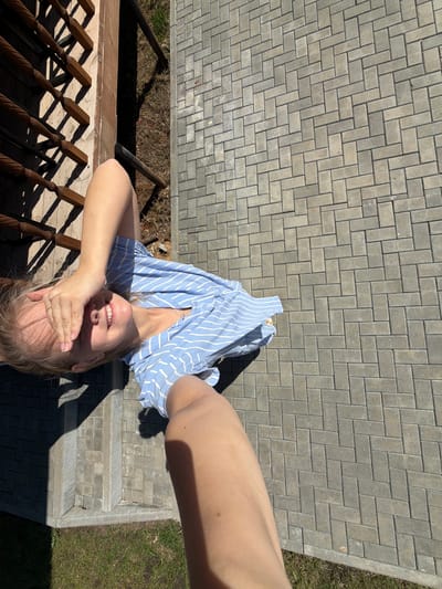 A woman in a blue and white striped top is lying on gray brick pavers, covering her eyes with her hand. Her head is near a wooden railing and dirt patch. A bare arm extends into the upper right corner of the image. The background includes brick pavers, a small grassy area, and part of a building foundation. There is no visible indication of the city of Kamennoye, Russia.