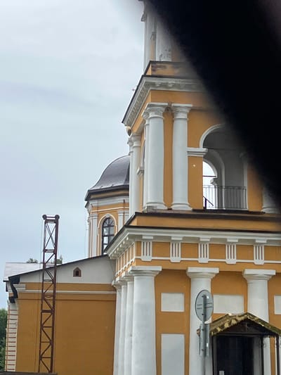 The image shows a portion of a yellow building with white columns and architectural details, suggesting it might be a historical or religious structure. A dark dome, likely part of the same building, is visible in the background. To the left, a tall, red, metal lattice structure stands against an overcast sky. A black, amorphous shape partially obscures the upper right portion of the image. The building exhibits signs of wear, with chipped paint visible on some of the upper sections. A small, covered entrance with a decorative roof and a signpost are visible at the bottom right. The overall impression is of an outdoor scene in a somewhat dilapidated urban or semi-urban environment.