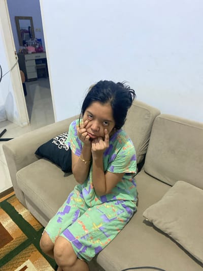 A young woman with dark hair and a green and purple patterned outfit sits on a beige sofa in a room. She is making a face, pulling down her eyelids with her fingers. A black pillow with a white swirl design is behind her to the left. To the right of the sofa is a tan pillow. In the background, a doorway leads to another room with a vanity table and a mirror. A rug with geometric patterns in brown, green, and white is on the floor in front of the sofa. The walls are white, and the lighting is artificial. The scene appears to be in a residential setting within Walennae, Indonesia.