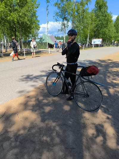 The image shows a man in black cycling gear and a helmet standing next to his bicycle on a sunny day. The bicycle is black with a red bag strapped to the back. In the background, there are trees, tents, and other people walking around. The ground is a mix of asphalt and dirt, with shadows from the trees cast on the ground. This scene is located in the city of Sertolovo, Russia.