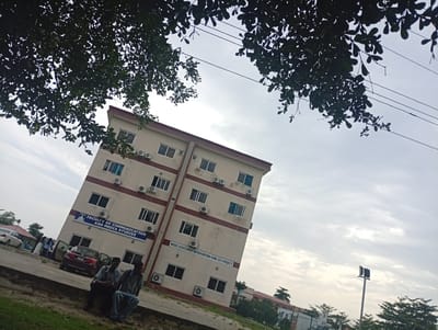 The image shows the Faculty of Communication and Media Studies building in Ijanikin, Nigeria. The building is a four-story beige structure with a red-brown trim. It has multiple windows and air conditioning units on each floor. A sign on the building reads "FACULTY OF COMMUNICATION AND MEDIA STUDIES" and "NEEDS ASSESSMENT INTERVENTION FUND 2013 PROJECT." Two men are sitting on a ledge in front of the building. Several cars are parked nearby and other people are in the vicinity of the building. Tree branches are visible in the foreground and top of the image. The sky is overcast. A light pole is in the background.