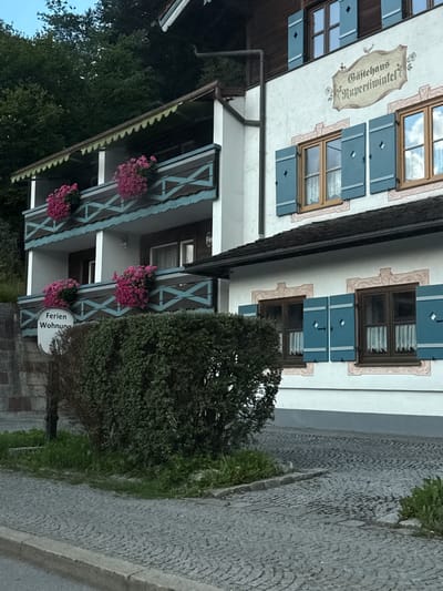 This is a full-frame, eye-level, outdoor, daytime, medium shot of a traditional Bavarian guesthouse named "Gasthaus Rupertwirt" located in Berchtesgaden, Germany. The building's facade is white with decorative trim around the windows and shutters. Blue shutters are a prominent feature on the windows. The guesthouse has a dark brown tiled roof and balconies adorned with pink flowers in planters. A sign on the lower balcony reads "Ferienwohnung", indicating holiday apartments. The building is situated on a cobbled driveway with a stone retaining wall and a dense green hedge in the foreground. Trees and foliage are visible in the background. There are no people or animals present in the image.