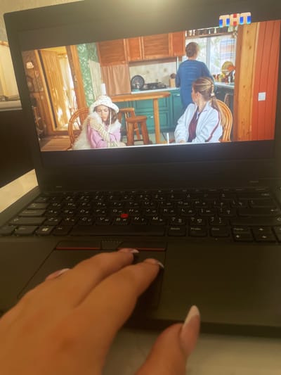 The image shows a laptop screen displaying a scene from a television show or movie. The scene appears to be set in a kitchen or dining area, with wooden cabinetry, a green countertop, and wooden stools. Three individuals are visible: a young person wearing a patterned hat and a fluffy white scarf, and two adults in the background. One adult is wearing a blue shirt and is standing, while the other is wearing a white jacket over a maroon top and is seated. A logo, possibly for a television channel, is visible in the top right corner of the screen. In the foreground, a person's hand is shown resting on the laptop's trackpad, with manicured fingernails visible. The laptop itself is black and has a Cyrillic keyboard layout. The image is taken from a slightly elevated angle, looking down at the laptop and the hand. The overall setting outside the frame is not visible.