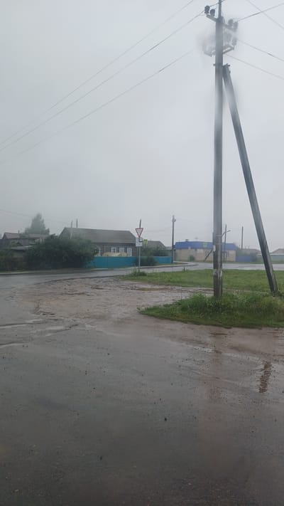 The image shows a street scene in the village of Kiyasovo, Russia, on a cloudy, overcast day, possibly with rain, as indicated by the wet pavement and reflections. In the foreground, a wet asphalt road curves to the left. To the right of the road is a patch of green grass and a power pole with multiple wires. In the middle ground, there is a cluster of wooden houses with dark roofs and a blue fence. A yield sign and a pedestrian crossing sign are visible near the road. Further in the background, more houses and trees are discernible through the hazy atmosphere. The overall impression is of a quiet, rural setting experiencing inclement weather.