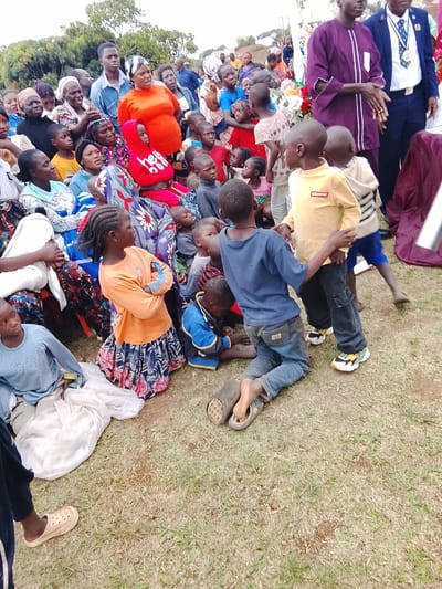 A gathering of people, including children, is taking place outdoors in Kuru, Nigeria. The individuals are diverse in age and attire, with some seated and others standing. Several children are playing or interacting near the front of the group. The setting appears to be a grassy area, possibly a park or open space, with trees visible in the background. Clothing styles are varied, with traditional African fabrics mixed with modern garments. Two men stand slightly apart from the larger group.