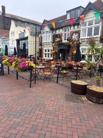 The image depicts the exterior of the Jolly Sailor pub located on Harbour Road, Poole, United Kingdom, on an overcast day. The pub's facade is visible, featuring a distinctive painted mural of a sailor. Colorful triangular flags are strung across the front of the building. Outdoor seating areas are arranged with numerous wooden tables and chairs, some covered by black umbrellas. Large planters filled with bright pink and yellow flowers are placed along a black metal railing in the foreground. Two large wooden barrels, used as planters filled with gravel, are situated on the brick-paved ground. The brick pavement features a herringbone pattern. A few patrons are seated at tables in the outdoor seating area. The overall scene suggests a commercial establishment preparing for or operating during outdoor service.