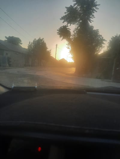 The image captures a street scene at sunrise or sunset in the city of Vosēābād, Tajikistan. The sun is low on the horizon, casting a warm, golden light that creates lens flare and silhouettes the trees and buildings. The view is from inside a vehicle, looking out through the windshield. The foreground shows the dark dashboard of the car with a small red indicator light visible. The middle ground displays a paved street lined with low-rise buildings, some with stone walls, and trees. Power lines are visible in the sky. The atmosphere is hazy, likely due to the low angle of the sun and possibly dust or mist. The environment appears to be a residential or rural area.