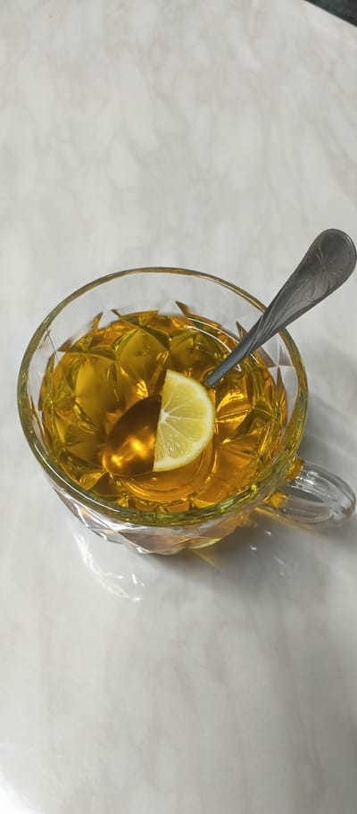 A close-up, top-down shot shows a glass mug filled with golden-yellow tea, a slice of lemon floating on the surface, and a silver spoon. The mug has a faceted design, and a handle is visible on the right side. The tea is clear and has a bright, warm hue, reflecting the light and creating patterns within the glass. The lemon slice is wedge-shaped, with visible pulp and rind. The spoon, resting partly in the tea and partly above it, features a decorative floral pattern on its handle. The setting appears to be indoors, with a light-colored, patterned surface beneath the mug, possibly a marble or granite countertop, as indicated by the lighting and subtle textures. The overall mood is one of simple refreshment, with the warm tones of the tea and the bright lemon suggesting a pleasant, perhaps morning or afternoon, beverage. There is no visible text in the image.
