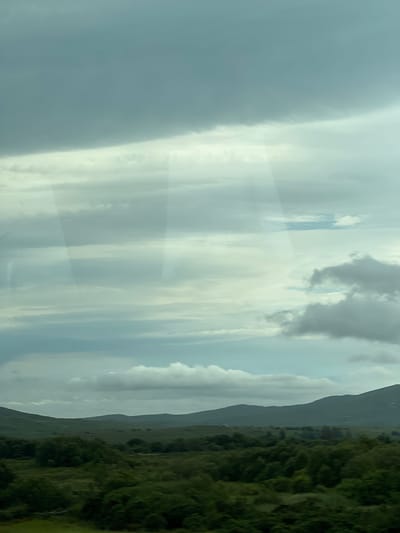 This media file captures a landscape scene, oriented vertically, from Drimnaraw, Ireland. The scene is dominated by a vast sky filled with dramatic clouds. On the left side, a large portion of the image is covered by a dense, grey cloud formation that appears to be either a thick cloud bank or perhaps a rain shower. This dark cloud contrasts with lighter, more diffuse cloud formations in the center of the image, which are streaked with pale yellow and white, suggesting shafts of light breaking through. The sky takes up the majority of the frame, indicating an expansive view.
To the right of the sky, a dark, undulating mountain range forms the middle ground. The mountains are mostly shrouded in shadow, with their contours softened by the distance and atmospheric conditions. Beyond the mountains, a vibrant, green landscape stretches out. This area is densely covered with vegetation, likely rolling hills or fields, characteristic of the Irish countryside. The greenery is rich and appears textured, with variations in shade indicating the uneven terrain and possibly the play of light and shadow.
The time of day is difficult to pinpoint precisely due to the cloud cover, but the presence of light suggests it is daytime, likely overcast or transitioning from rain. The weather appears to be cloudy, possibly with recent or impending rain given the heavy clouds on the left. The location cues – the lush green vegetation and the mountainous terrain – are consistent with the setting of Drimnaraw, Ireland. There are no visible people or objects, and no text is present in the image. The overall mood is one of natural grandeur and perhaps a touch of dramatic weather.