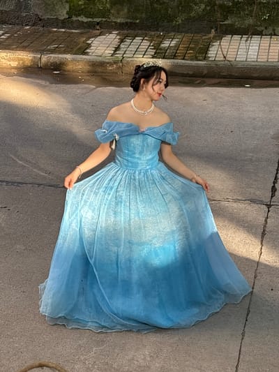 A woman in a light blue gown lies on a concrete surface. She wears a tiara and a pearl necklace. The concrete is likely a pathway and is near a stone wall covered in vegetation. The image was possibly taken in Baqiao, China.