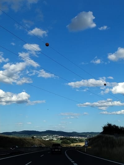 The image shows a bright blue sky with scattered white clouds. Several power lines stretch across the frame. Two dark, spherical objects, possibly weights or markers, hang from the power lines. In the distance, a landscape with rolling hills, fields, and a town is visible under a hazy sky. A highway is in the foreground, with cars driving on it. The scene appears to be a rural or semi-rural area, possibly on the outskirts of Mladotice, Czechia, given the context. The overall impression is one of a sunny day with a highway view.