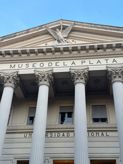 The image depicts the facade of the Museo de La Plata (La Plata Museum) in La Plata, Argentina. The perspective is from below, looking upwards at an angle. The architectural details prominently displayed include neoclassical columns with ornate Corinthian capitals. Inscribed vertically on the building's facade is the text "MUSEO DE LA PLATA" and "UNIVERSIDAD NACIONAL." A sculptural element depicting a winged figure is positioned on the pediment. The sky is visible in the upper left corner. Windows are visible between the columns.