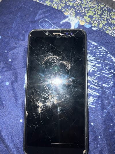 A smartphone with a severely cracked screen lies horizontally on a patterned fabric. The screen's damage consists of multiple radial cracks, creating a web-like appearance, particularly around the center. The phone's bezel is visible along the edges, and some debris is trapped in the cracks. The fabric background appears to have a blend of blue, purple, and white patterns, with some floral elements visible near the top left.