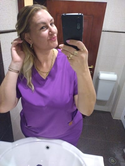 A woman is taking a selfie in a bathroom. She is wearing a purple scrub top and gold jewelry. She has blonde hair and is looking at the camera with a smile. The bathroom has white tiled walls and a dark tiled floor. A white trash can is mounted on the wall. The image was taken in Yagoda, Bulgaria.