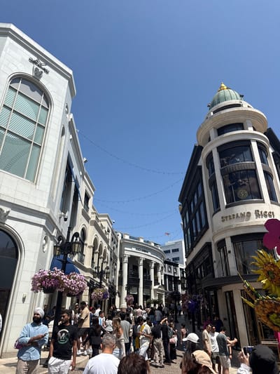 The image captures a bustling scene in Beverly Hills, California, United States. The street is lined with high-end retail buildings, predominantly white with ornate architectural details. People are walking along the street, indicating a lively shopping environment. Visible storefronts and building facades suggest a mix of designer boutiques and commercial establishments. Overhead, decorative string lights are strung across the street. The sky is clear blue, suggesting a sunny day. A few decorative planters and flower arrangements add to the visual aesthetic of the area.