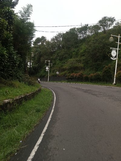 The image displays a curving asphalt road through a hilly, vegetated area in Deothang, Bhutan, under an overcast sky. The road, which features a solid white line on its right edge and a less distinct dashed center line, curves to the left from the foreground. On the immediate left, a stone retaining wall supports an embankment covered in green grass and dense foliage. Further left and along the hillside, tall green trees dominate. On the right side of the road, several white street lamp posts with decorative, circular cream-colored signs and lanterns are spaced along the curve. The hillside to the right is also densely covered with green trees and smaller reddish-brown bushes. A single individual on a dark motorcycle, wearing a white shirt, is visible in the distance on the left side of the road, navigating the curve away from the viewer. Overhead power lines are visible against the sky in the upper middle portion of the image.