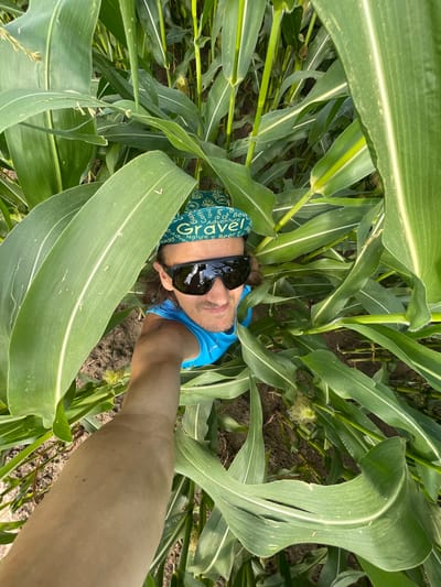 A high-angle, eye-level selfie shows a person in a bright blue tank top and a green bandana with the word "Gravel" prominently displayed. The person is wearing dark sunglasses and has medium-length brown hair. They are positioned within a dense field of tall green corn stalks, with large leaves partially obscuring their face and upper body. The background consists of more corn plants and dark soil. The lighting suggests it is daytime. The image was taken in Perevoznoe, Russia.